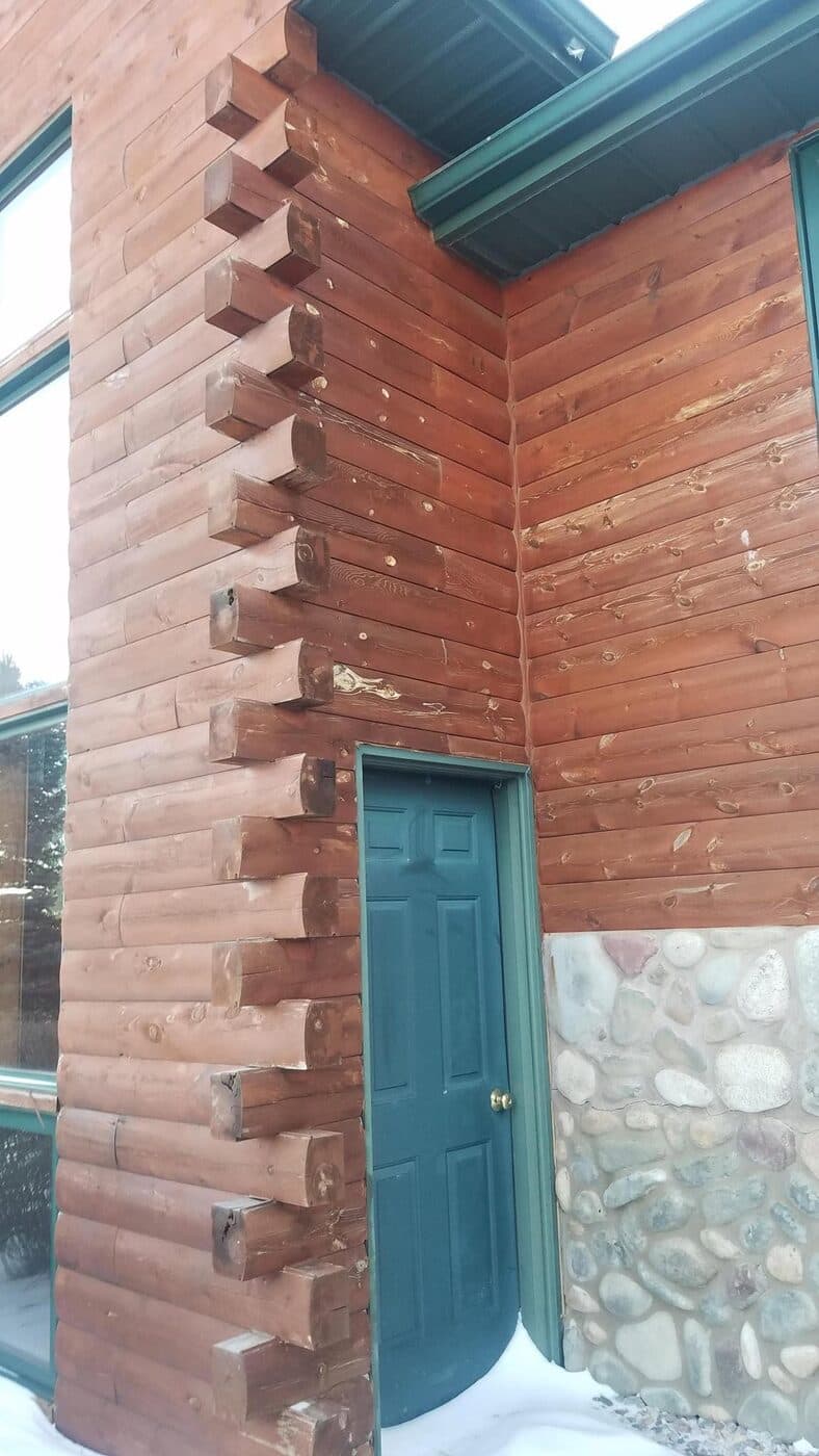 A saddle-notched log-siding corner in warm stained cedar over a river-rock stone base, with a green door and green metal fascia.