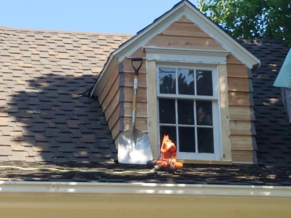 A re-roof in progress up close: brown architectural shingles laid tight around a dormer, with a roofing gun and a shovel resting on the deck.