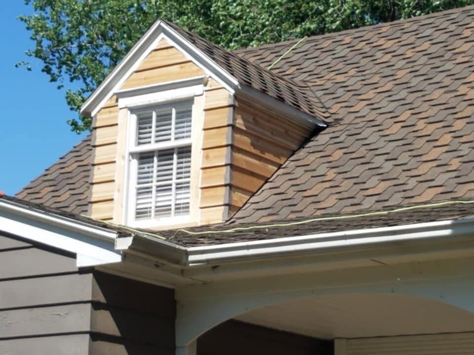 A steep gable dormer clad in fresh cedar, wrapped in new brown architectural shingles under a clear blue sky.