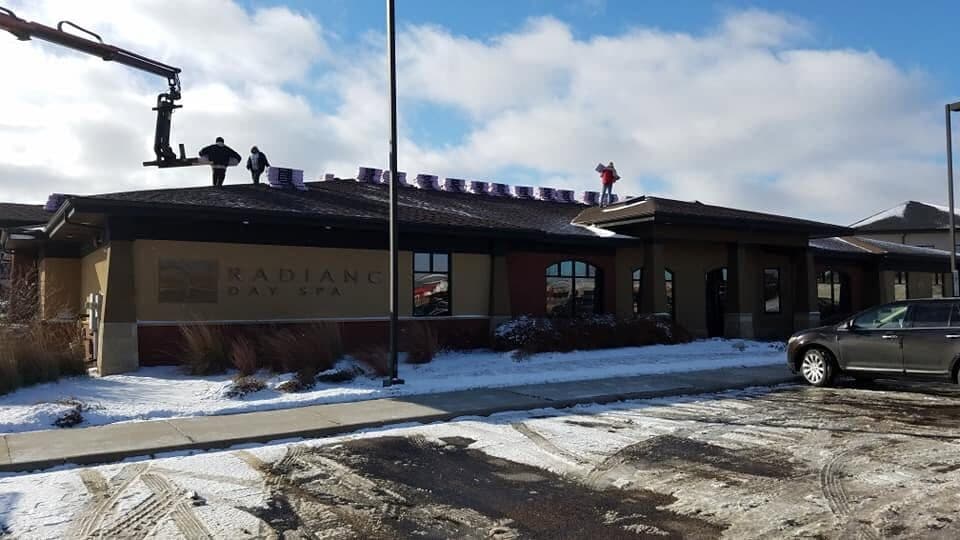 A winter commercial re-roof: shingle bundles staged along the ridge of a single-story building while a boom lift feeds material to the roof.