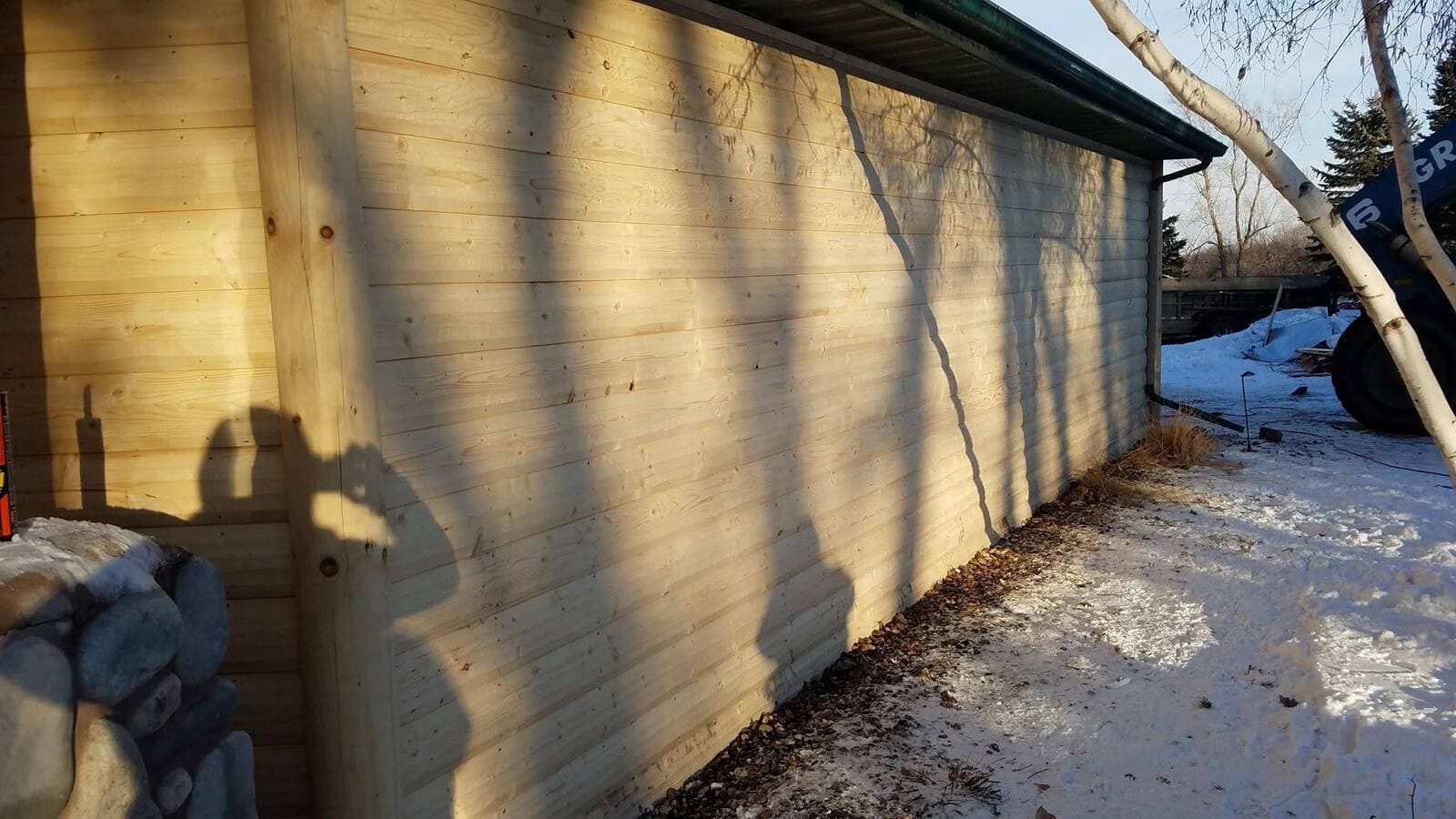Fresh natural-wood log-lap siding on an outbuilding at golden hour, river-rock stone at the corner and green metal fascia above.