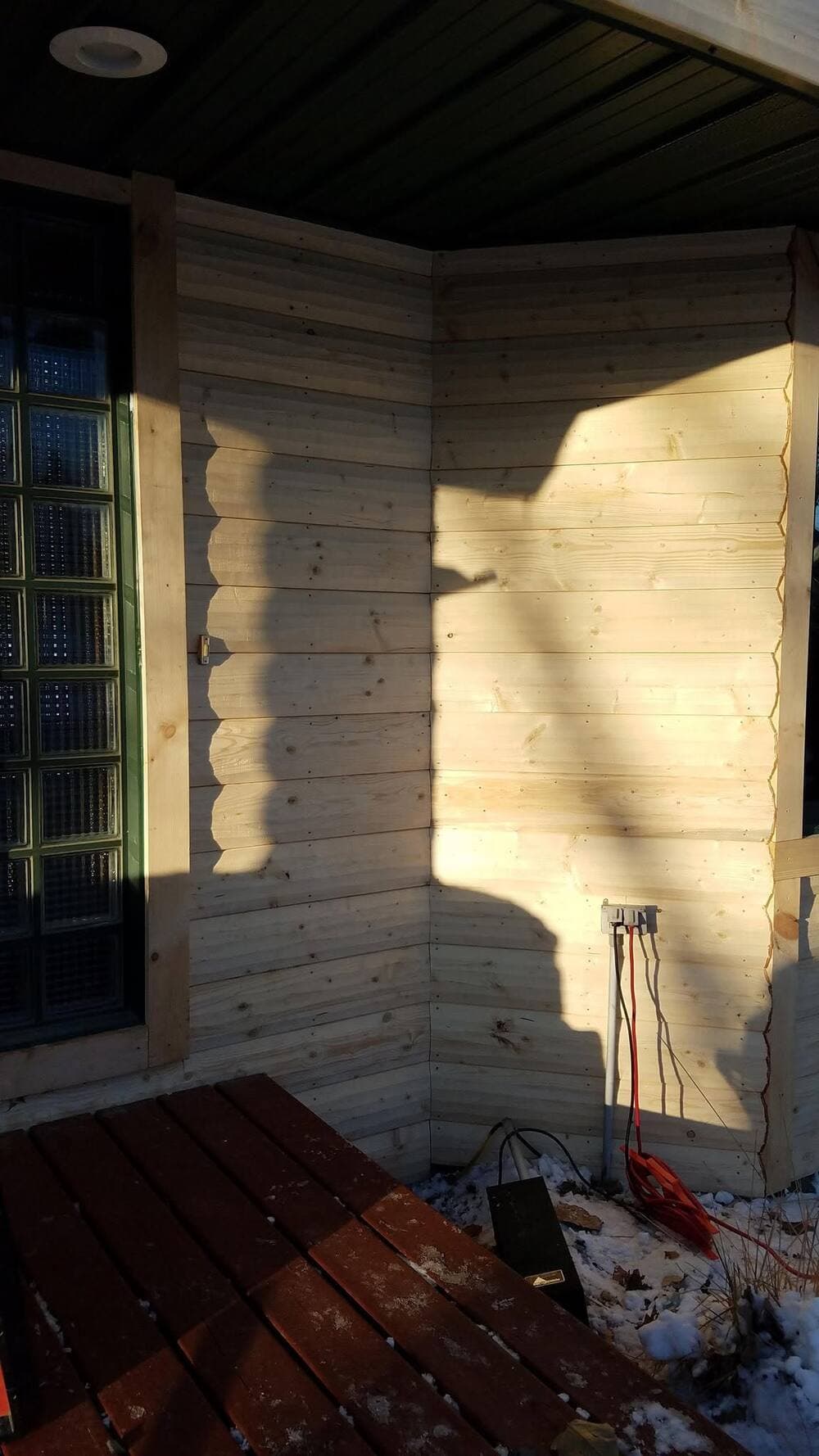 A mitered corner of fresh natural-wood log-lap siding under a green metal soffit, with a glass-block sidelight and a green-trimmed door.