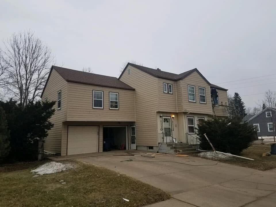 A two-story duplex mid re-side, fresh tan lap climbing the walls with the old materials cleared to the driveway.