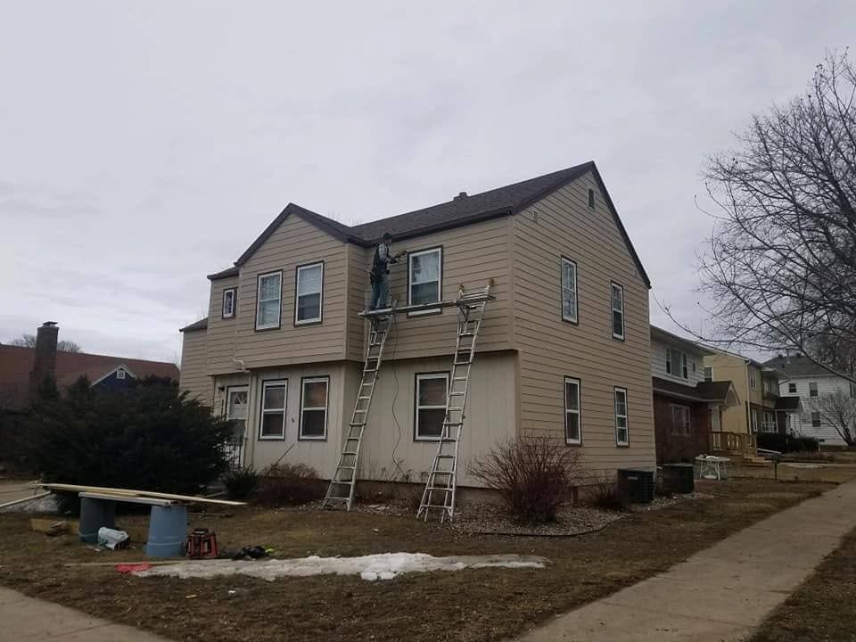 A Specialty crew member re-siding the second story of a home from an extension ladder, fresh greige lap going up over the old.