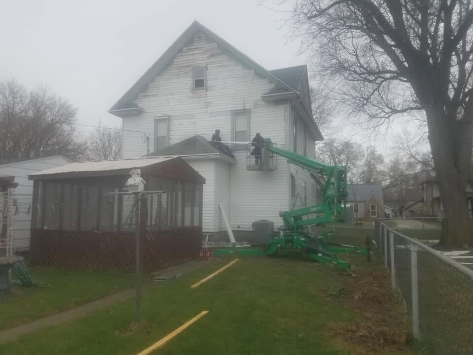 A two-story farmhouse re-side underway on a gray day, the crew working the upper walls from a green articulating boom lift.