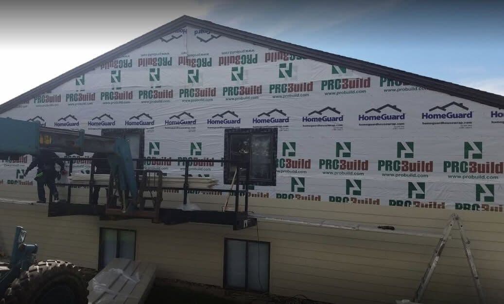 A full gable wrapped in HomeGuard house-wrap with new lap siding started along the base, a crew member setting boards from a lift platform.