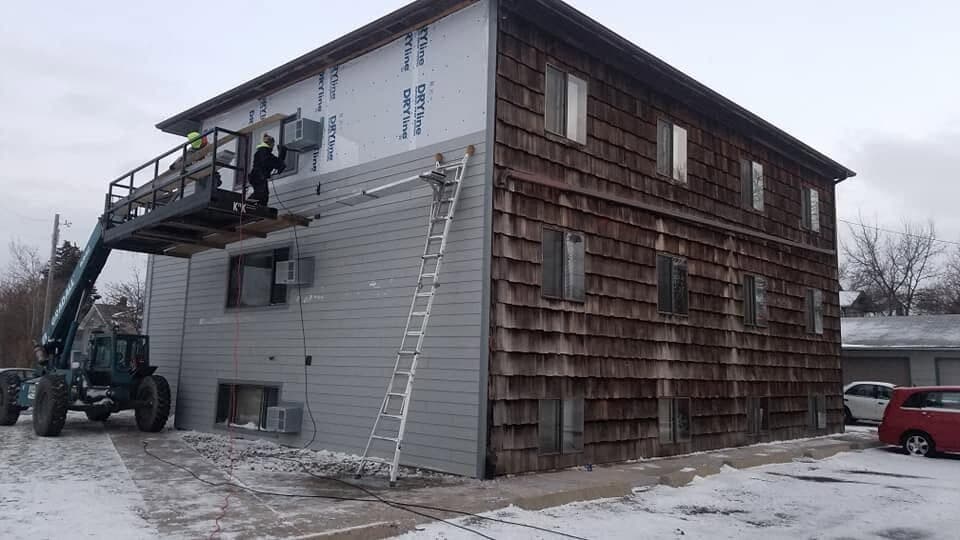 A multi-story re-side in progress: new lap siding over house-wrap from a telehandler platform, beside the existing cedar shake.